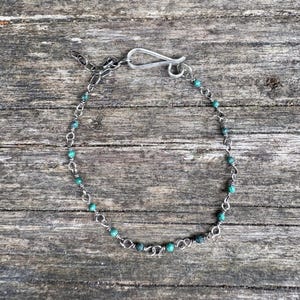 May include: A silver chain bracelet with small, round, teal beads. The bracelet has a hook clasp and an adjustable chain. The teal beads are evenly spaced along the silver chain. The bracelet is approximately 18 cm in length.