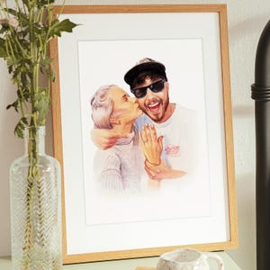 May include: A framed watercolor portrait of a couple, with the woman kissing the man on the cheek. The man wears sunglasses and a baseball cap. The frame is light brown, and the artwork is displayed on a white background.