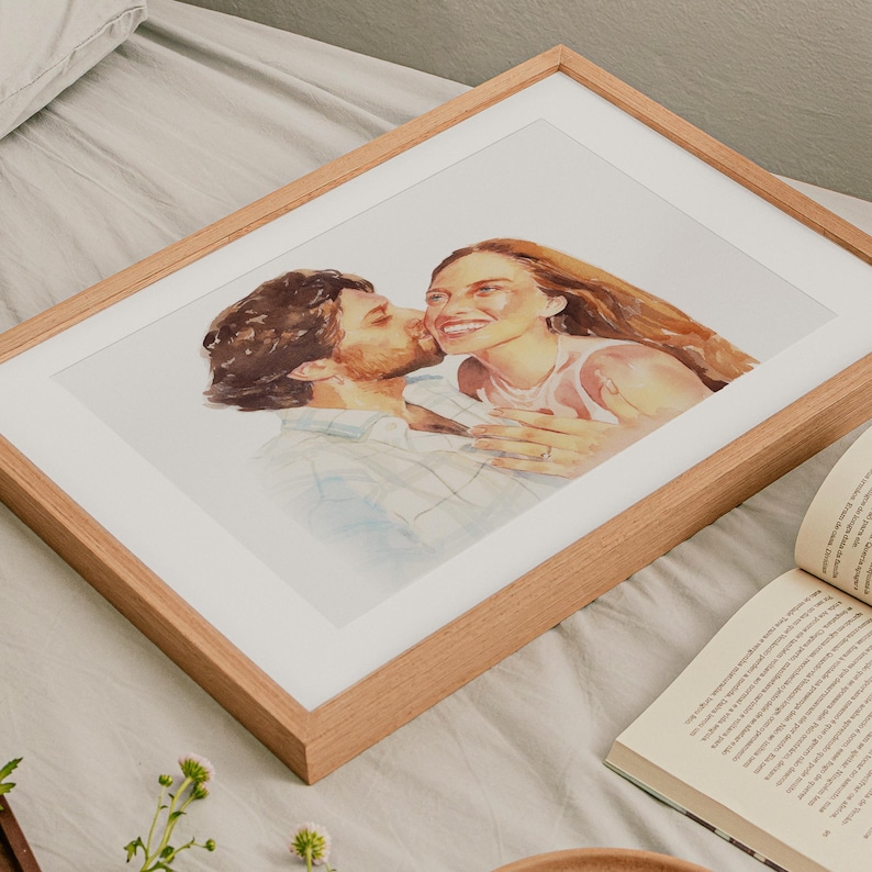 May include: A framed watercolor portrait of a couple, with the man kissing the woman's cheek. The artwork is in a light wood frame with a white mat. The woman has long, flowing auburn hair and is smiling. The man has dark hair and a beard.