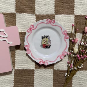 May include: A pink cross-shaped wooden block with a white bow, a white ceramic plate featuring a painted pink ribbon and bow border with a central Dolly Parton 9 to 5 pin, and a decorative branch with small pink and white heart-shaped accents.
