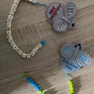 Op de afbeelding: Fopspeenclips met houten alfabetkralen en muisvormige applicaties. Eén clip heeft blauwe en groene kralen, de andere witte alfabetkralen. De applicaties zeggen "Schulkind 2024".