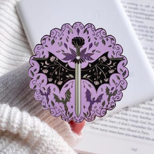 May include: A circular enamel pin featuring a central silver sword with a dark flower hilt, flanked by black bat wings with floral details. The background is light purple with a scalloped border and small dragon silhouettes in purple and green.