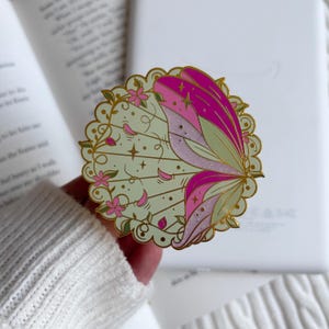May include: A circular enamel pin featuring a pale green background with gold scalloped edges. The design displays a stylised wing in shades of bright pink and light lavender with glitter accents, surrounded by small pink flowers and gold star motifs.