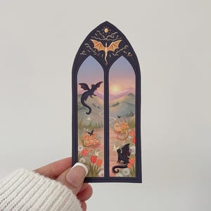 May include: A sticker featuring a gothic window design with dragons. The window frame is dark blue, and the scene inside depicts dragons in flight and resting in a field of tulips. The sky is a gradient of orange and blue.