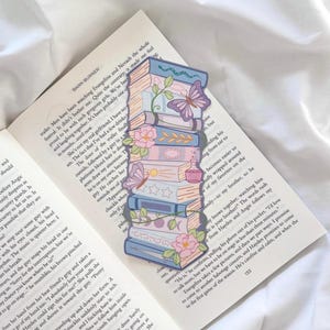 May include: A decorative bookmark depicting a stack of books with floral and butterfly details. The bookmark uses pastel colours, including blue, pink, and purple. It rests on the open pages of a book.