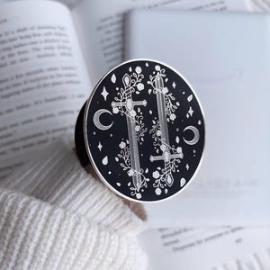 May include: A round, black and silver pin featuring two crossed swords, floral accents, stars, and crescent moons. The pin is set against a backdrop of an open book and a white sweater.
