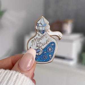May include: A white and gold enamel pin in the shape of a potion bottle. The bottle contains blue glitter and the words "Moon Dust". The top of the bottle is a faceted crystal shape.