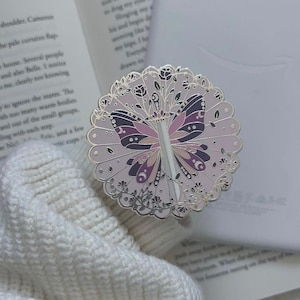 May include: A round, pink enamel pin featuring a butterfly design with a sword through its center. The butterfly has purple and pink wings, and the pin is surrounded by floral and leaf details. The pin is set against an open book and a white sweater.