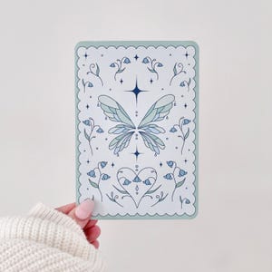 May include: A rectangular card with a scalloped edge featuring a central illustration of light blue butterfly wings, surrounded by delicate line art of small bell-shaped flowers, stars, and a heart shape, all set against a white background.