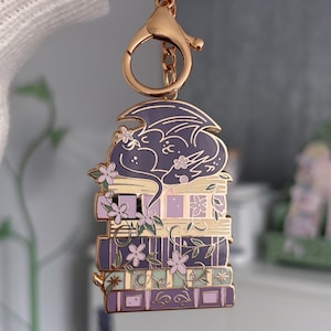 May include: A gold-coloured keychain featuring a purple dragon resting on a stack of books. The books are in shades of purple, lavender, and green, with floral details. The keychain has a lobster clasp.