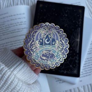 May include: A circular enamel pin with a scalloped gold edge featuring a purple and blue illustration of a dragon sleeping in a garden under a crescent moon and stars, framed by an ornate archway with wings and small floral accents.