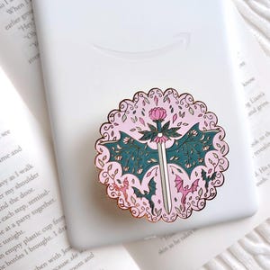 May include: A circular phone grip featuring a rose gold scalloped edge and a central design of a silver sword topped with a pink flower. The background is light pink with dark teal bat wings, small dragons, and floral vines in a decorative pattern.