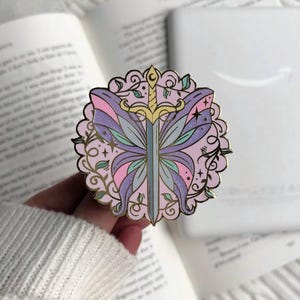 May include: A pastel enamel pin with a butterfly and sword design. The wings are purple, pink, and blue, with gold detailing. The pin has a gold filigree border and small green leaves. The pin is approximately 4 cm in diameter.