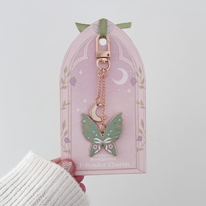 May include: A rose gold metal charm featuring a green butterfly with white and pink accents and a small crescent moon, attached to a chain with a lobster clasp. The charm is displayed on a light pink card labeled E-Reader Charm.