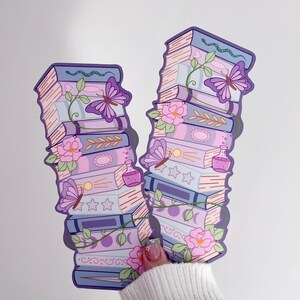 May include: Two vertical bookmarks featuring stacked books in shades of purple, blue, and pink. The illustrations include purple butterflies, green vines, pink flowers, small potion bottles, and moon phase symbols printed on the book spines.