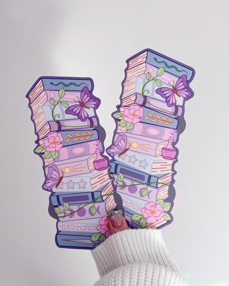 May include: Two vertical bookmarks featuring stacked books in shades of purple, blue, and pink. The illustrations include purple butterflies, green vines, pink flowers, small potion bottles, and moon phase symbols printed on the book spines.