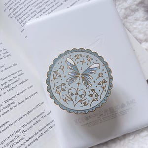 May include: A circular phone grip with a scalloped gold edge, featuring a light blue background, a central butterfly illustration, and a heart motif surrounded by small floral sprigs. The accessory is attached to a white e-reader device.