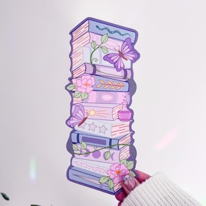 May include: A vertical bookmark featuring a stack of illustrated books in shades of purple, lavender, and pink. The design includes a butterfly, a snake, pink flowers, a small potion bottle, and moon phases, all outlined in a dark purple border.