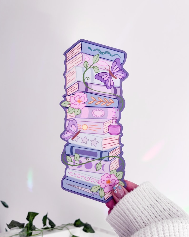May include: A vertical bookmark featuring a stack of illustrated books in shades of purple, lavender, and pink. The design includes a butterfly, a snake, pink flowers, a small potion bottle, and moon phases, all outlined in a dark purple border.