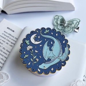 May include: A round enamel pin with a scalloped gold edge featuring a dark blue background, a light blue dragon, a white crescent moon, small white flowers, and tiny butterflies. The dragon and butterflies have a subtle glittery finish.