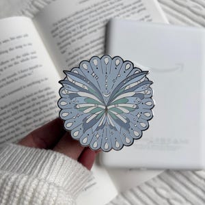 May include: A circular enamel pin featuring a symmetrical butterfly design in shades of light blue, teal, and white. The butterfly is surrounded by a scalloped border with teardrop shapes and small star accents, all outlined in a dark metal finish.