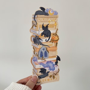 May include: A whimsical sticker featuring a stack of books with a fantasy theme. The books are adorned with small black dragons, a crescent moon, stars, and other magical elements. The color palette includes beige, blue, and gold.