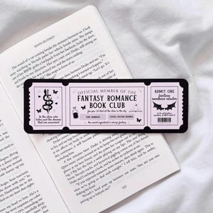 May include: A pink and black bookmark with a ticket design. The bookmark features a black and white illustration of a bat and the text "Admit One Fantasy Romance Reader". The bookmark also features a black and white illustration of a potion bottle and the text "Official Member of the Fantasy Romance Book Club".