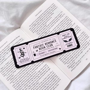 May include: A black and pink bookmark with a ticket design. The bookmark features a pink background with black text that reads "Official Member of the Fantasy Romance Book Club". The bookmark also features a black and white illustration of a bat with the text "Admit One Fantasy Romance Reader".