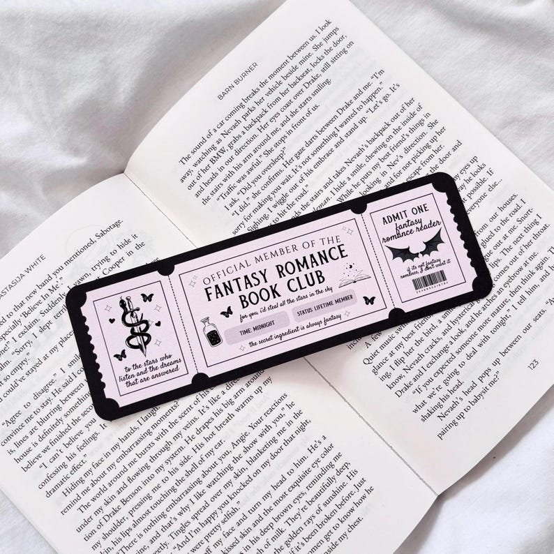 May include: A black and pink bookmark with a ticket design. The bookmark features a pink background with black text that reads "Official Member of the Fantasy Romance Book Club". The bookmark also features a black and white illustration of a bat with the text "Admit One Fantasy Romance Reader".
