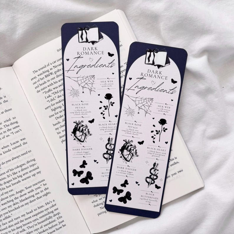 May include: Two dark blue bookmarks with white text that reads "Dark Romance Ingredients". Each bookmark features a black and white illustration of a heart, rose, dagger, spider web, and butterflies.