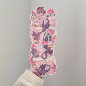 May include: A sticker depicting a stack of books adorned with pink flowers and purple dragons. The design includes a shield and an arched window, all in a fantasy theme.