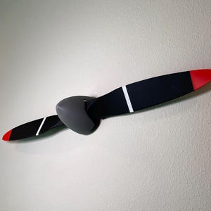 May include: A black and white airplane propeller with red tips mounted on a gray wall.