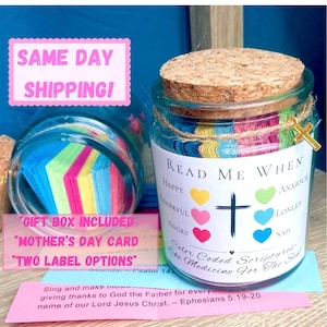 May include: A glass jar with a cork lid, filled with colorful paper strips. The jar has a label that reads "Read Me When" with various emotions listed. A small gold cross charm hangs from the jar. The jar is a unique gift idea.