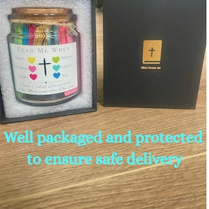 May include: A glass jar filled with colorful paper strips, labeled "Read Me When" with emotions like happy and sad. A cross charm hangs from the lid. The jar is in a black box with a gold cross.