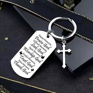 May include: A silver-toned metal keychain featuring a rectangular dog tag pendant and a small cross charm. The tag is engraved with the text: Happy Moments, Praise God. Difficult Moments, Seek God. Quiet Moments, Worship God. Painful Moments, Trust God. Every Moment, Thank God.