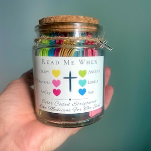 May include: A glass jar with a cork lid and a cross charm, filled with colorful paper strips. The jar has a label that reads "Read Me When" followed by various emotions. The text "Color Coded Scriptures - Like Medicine For The Soul" is also visible.