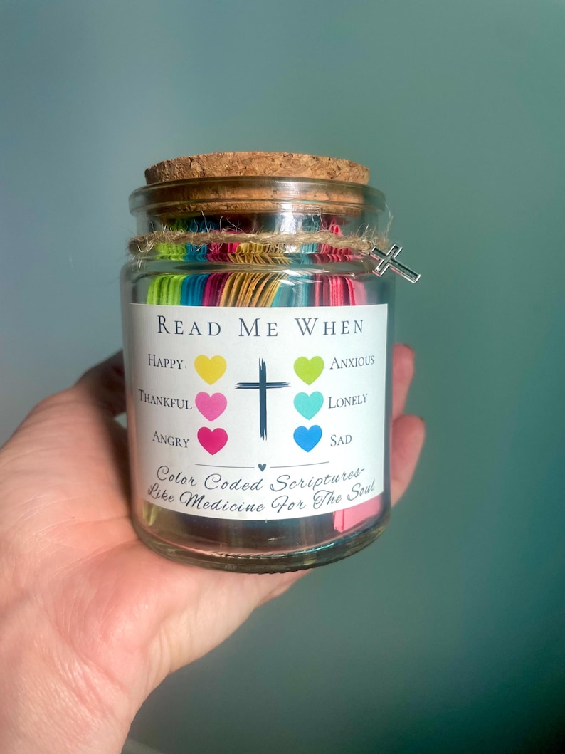 May include: A glass jar with a cork lid and a cross charm, filled with colorful paper strips. The jar has a label that reads "Read Me When" followed by various emotions. The text "Color Coded Scriptures - Like Medicine For The Soul" is also visible.
