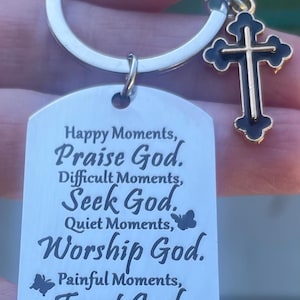 May include: A silver-colored keychain with a rectangular tag and a cross charm. The tag has the text "Happy Moments, Praise God. Difficult Moments, Seek God. Quiet Moments, Worship God. Painful Moments, Trust God. Every Moment, Thank God."