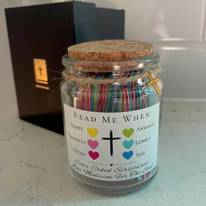 May include: A glass jar with a cork lid, filled with colorful paper strips. The jar has a label that reads "Read Me When" with various emotions listed. A gold cross charm hangs from the twine around the jar. A black box is in the background.