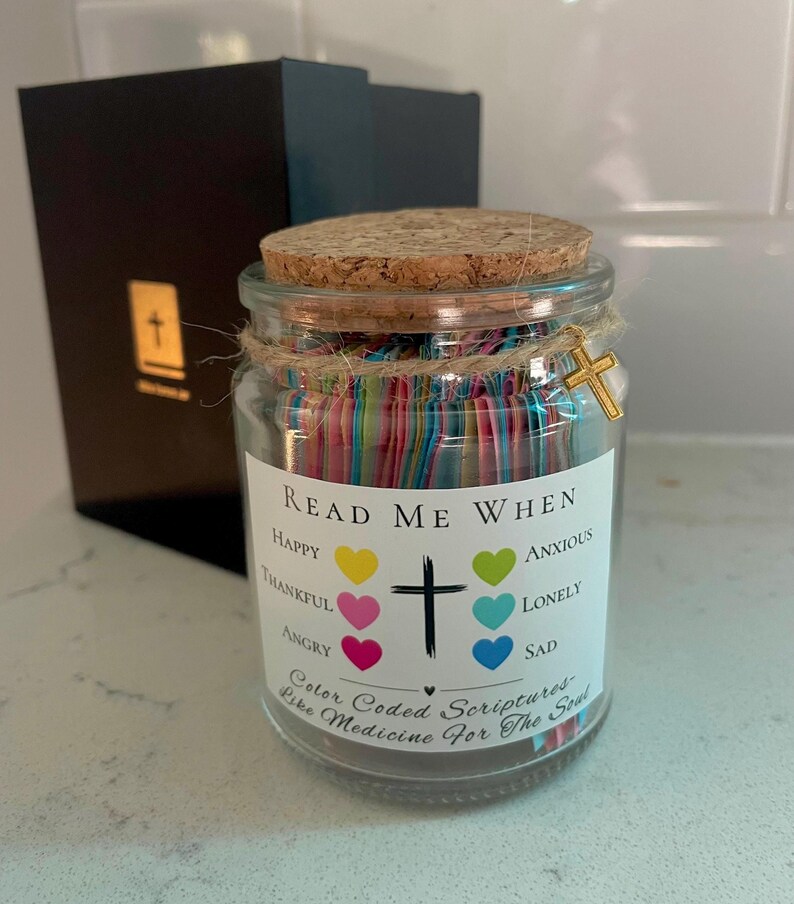 May include: A glass jar with a cork lid, filled with colorful paper strips. The jar has a label that reads "Read Me When" with various emotions listed. A gold cross charm hangs from the twine around the jar. A black box is in the background.