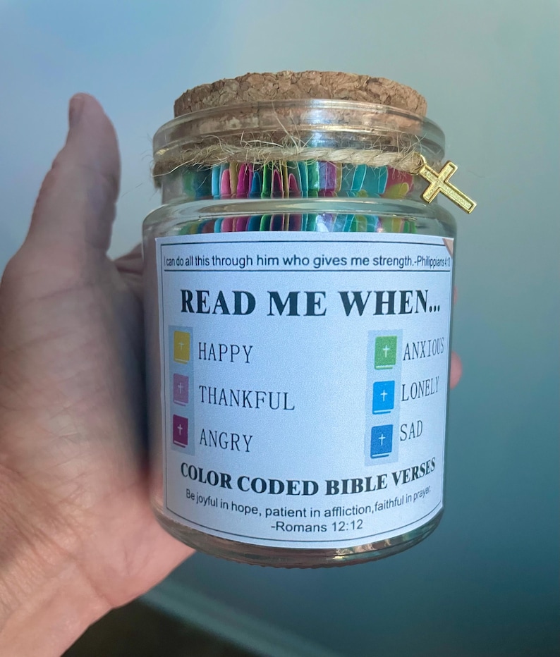 May include: A glass jar with a cork lid and a gold cross charm. The jar contains colorful paper strips and a label that reads "Read Me When..." with prompts for different emotions and Bible verses. The jar is held by a person.