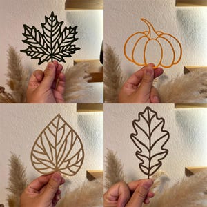 Può includere: Quattro ritagli a tema autunnale: una foglia d'acero nera, una zucca arancione, una foglia beige e una foglia di quercia marrone. Ognuno è tenuto da una mano su uno sfondo neutro, suggerendo decorazioni stagionali o forniture artigianali.