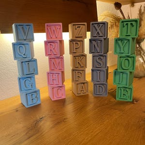 Op de afbeelding: Gestapelde alfabetblokken in verschillende pastelkleuren, waaronder blauw, roze, beige, grijs en groen. Elke blok toont een andere letter van het alfabet. De blokken zijn in verticale kolommen gerangschikt, wat een speelse en educatieve weergave creëert.