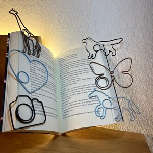May include: A collection of wire bookmarks in various shapes, including a giraffe, a heart, a camera, a dog, a butterfly, and a horse. The bookmarks are placed in an open book, with the giraffe bookmark at the top.