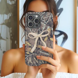 May include: A phone case with a camouflage pattern, featuring a cream-colored bow and the name "ANN" in a serif font. The case has a multi-lens camera system.
