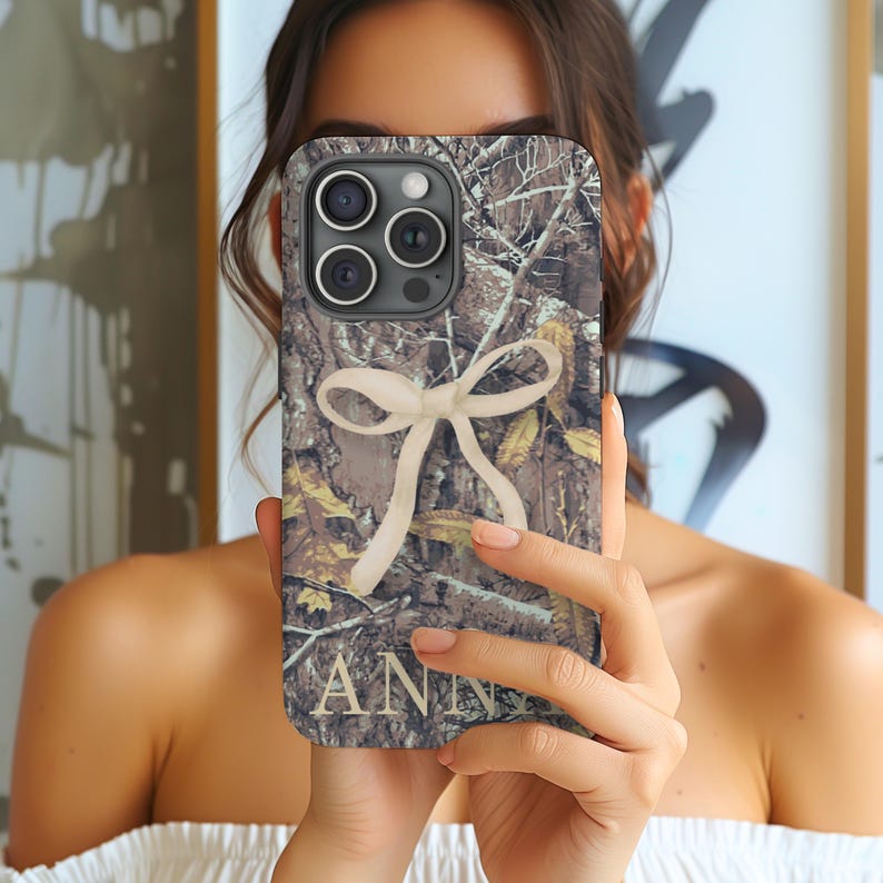 May include: A phone case with a camouflage pattern, featuring a cream-colored bow and the name "ANN" in a serif font. The case has a multi-lens camera system.