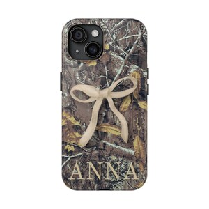 May include: A phone case featuring a camouflage pattern with a cream-colored bow design. The name "ANNA" is printed in a serif font at the bottom of the case. The case has a black border and a camera cutout.
