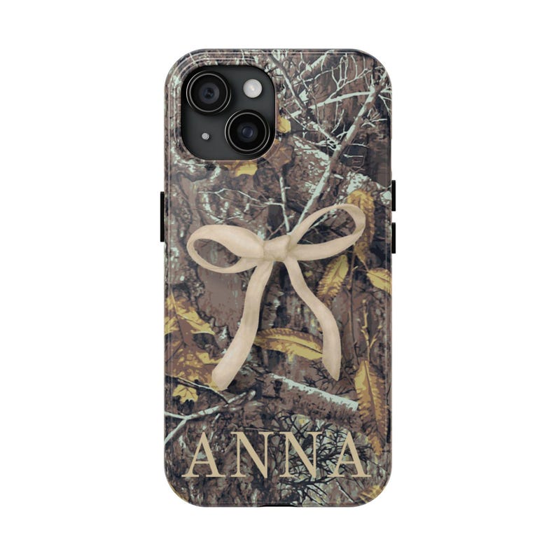 May include: A phone case featuring a camouflage pattern with a cream-colored bow design. The name "ANNA" is printed in a serif font at the bottom of the case. The case has a black border and a camera cutout.