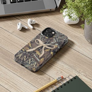 May include: A camouflage-patterned phone case with the name "ANNA" in gold lettering. A beige bow design is centered on the case. The phone case is on a wooden surface, with a laptop, earbuds, and a small plant in the background.