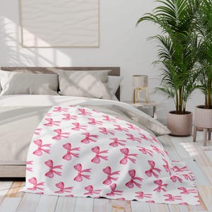 May include: A white blanket featuring a repeating pattern of pink watercolour bows. The bows are evenly spaced across the blanket's surface. The blanket is draped over a bed with neutral-coloured bedding.